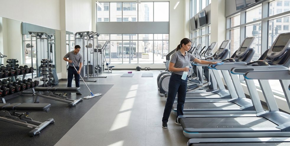 Cleaners disinfecting exercise equipment in a modern fitness center with treadmills and weight benches